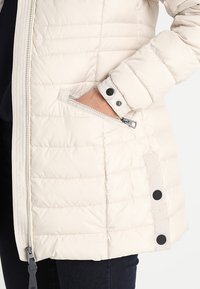 Lys beige pufferjakke med vandret quiltet design, lynlåslomme og sorte trykknapdetaljer på siden, med en glat tekstur.