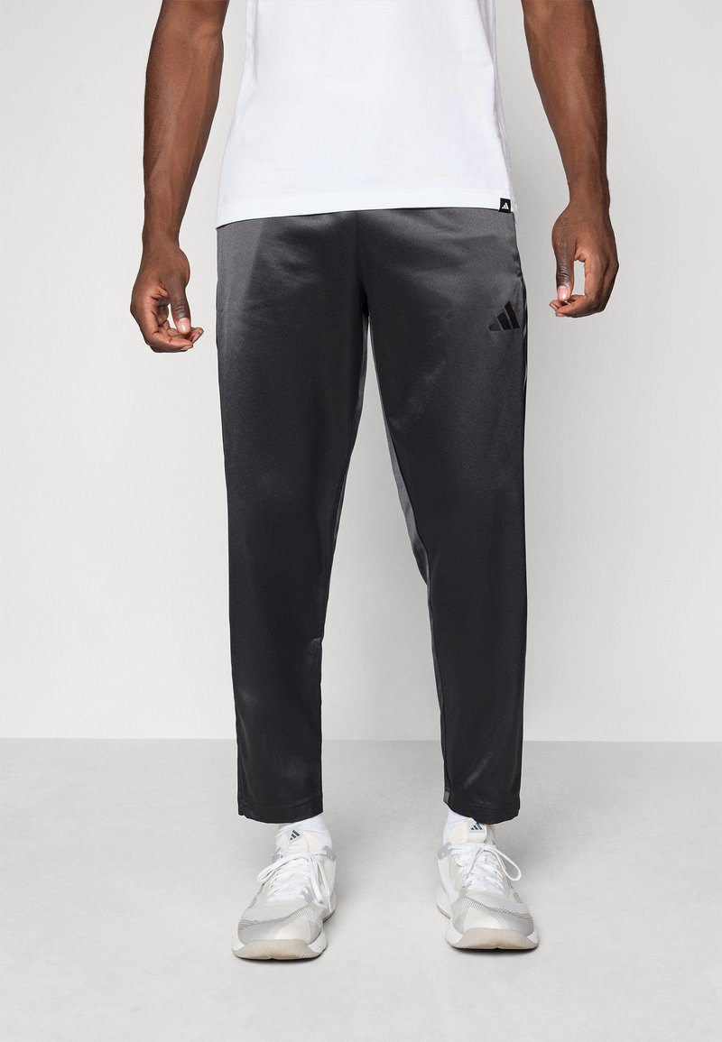 Sorte, skinnende atletiske bukser med slim fit, der har et Adidas-logo på venstre hofte, parret med lysegrå sneakers og hvide sokker.