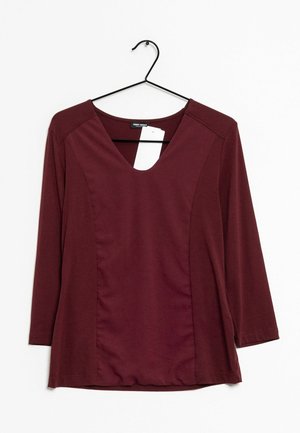 Bordowy top z długim rękawem i dekoltem w kształcie litery V, gładki materiał oraz boczne panele. Metka widoczna w pobliżu dekoltu, zawieszony na czarnym plastikowym wieszaku.