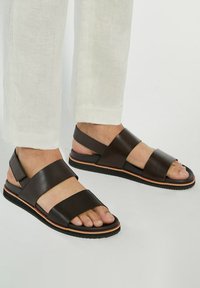 Brown leather sandals featuring wide straps, open toe design, and a textured black sole with an orange accent band.