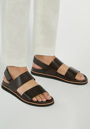 Brown leather sandals featuring wide straps, open toe design, and a textured black sole with an orange accent band.