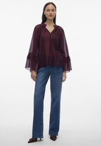 Frau trägt eine transparente Pflaumenbluse mit gerafften Glockenärmeln, blaue Jeans mit geradem Bein und spitze braune Schuhe vor weißem Hintergrund.