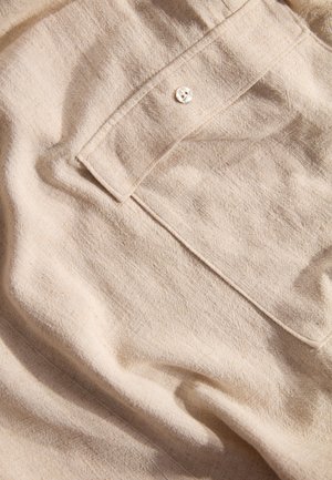 Helles beiges Gewebe mit einer strukturierten Oberfläche, ausgestattet mit einem einzigen Knopfverschluss und einer Tasche, vermittelt das Aussehen von natürlichem Leinen.