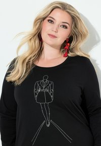 Blondýnka s rozpuštěnými vlasy má na sobě černý top s designem modelky na přehlídkovém molu zdobeným kamínky a velké červené výrazné náušnice, dívá se do kamery.