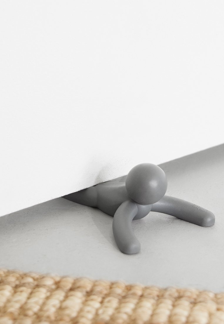 Grauer Türstopper in Form einer kleinen Figur mit runder Kopfform und ausgestreckten Armen, zwischen einer Tür und dem Boden auf einer hellen Oberfläche platziert.