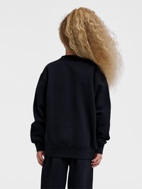 Dziecko z długimi kręconymi blond włosami, ubrane w luźną czarną bluzę i pasujące spodnie, stoi tyłem na jednolitym tle.