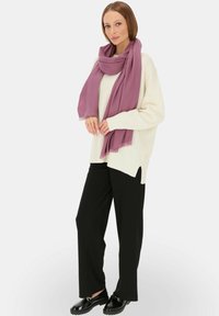 Przesadnie duży, biały sweter dzianinowy, luźne czarne spodnie oraz długi lawendowy szalik z postrzępionymi brzegami. Modelka pozuje z rękami umieszczonymi na szaliku.