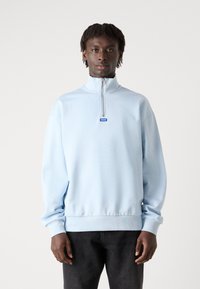 Sudadera de medio cierre en azul claro con puños y dobladillo acanalados, que presenta un pequeño parche de logo azul en el pecho, fabricada con un material suave y liso.