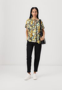 Blomstret bluse med gule, oransje og teal trykk på en mørk bakgrunn; løs passform med en nøkkelhulopening ved halsen. Sorte bukser og hvite sneakers.