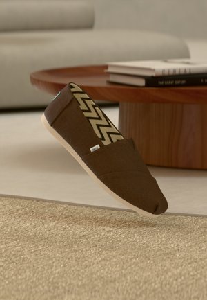 Chaussure marron à enfiler avec une doublure intérieure à motif zigzag, placée près d'une table basse ronde en bois avec des livres empilés dans un salon.