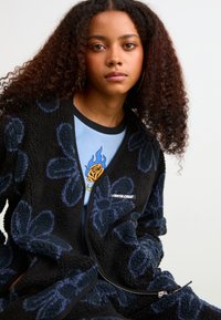 Giacca in pile nera con motivo floreale blu, dotata di una cerniera frontale e del logo 'Santa Cruz'. Indossata sopra una camicia blu chiaro con una grafica di rosa.