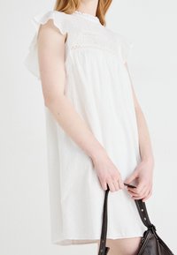 Valkoinen hihaton mekko, jossa on röyhelöhihoja, kierrätetty kuosi ja monimutkainen smokkikuvio kaula-aukossa. Malli pitää tummasta käsilaukusta kiinni.