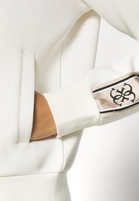 Sudadera color crema con puños acanalados, con un bolsillo lateral y un logo bordado en un panel de acento. Textura de tela suave.