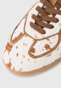 Sneaker featuring a white and brown spotted upper with a textured finish, brown laces, and a gum rubber sole. Unique design elements noted.