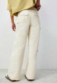 Pantaloni cargo cu croială largă din țesătură bej moale, având două buzunare la spate cu închidere prin buton și o textură fină, ușoară.