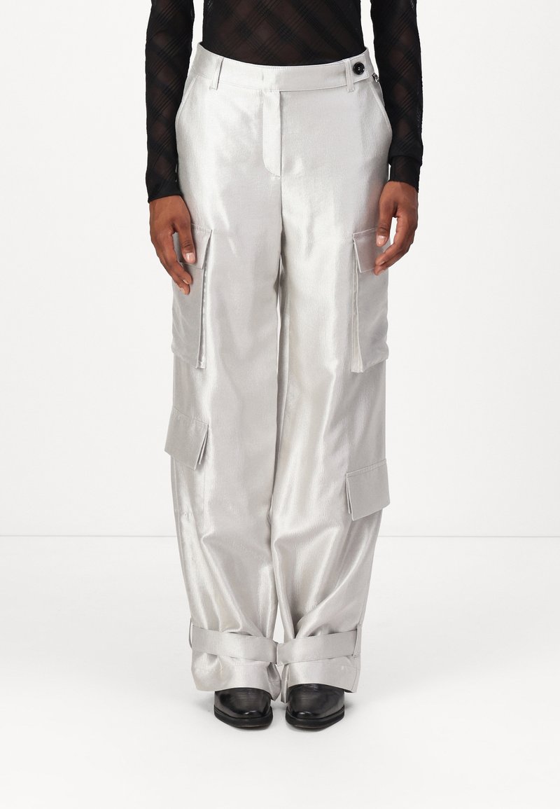 Personne portant un pantalon cargo argenté brillant avec plusieurs poches et des sangles aux chevilles, associé à des chaussures noires et à un haut noir à manches longues.