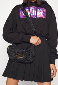 Černá nařasená nylonová taška se zlatou logovou destičkou, obsahující ramenní popruh a zipovou kapsu. Modelka má na sobě černou mikinu a plisovanou sukni.