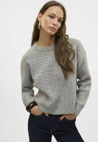 Suéter de lana gris con textura acanalada, cuello redondo y mangas largas, combinado con jeans de mezclilla oscura y una pulsera de madera.