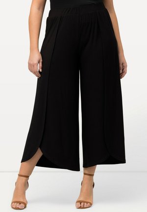 Pantalon noir large et court aux jambes larges avec des panneaux frontaux superposés, porté avec des talons marron à bride à la cheville, vu de la taille jusqu'aux pieds.