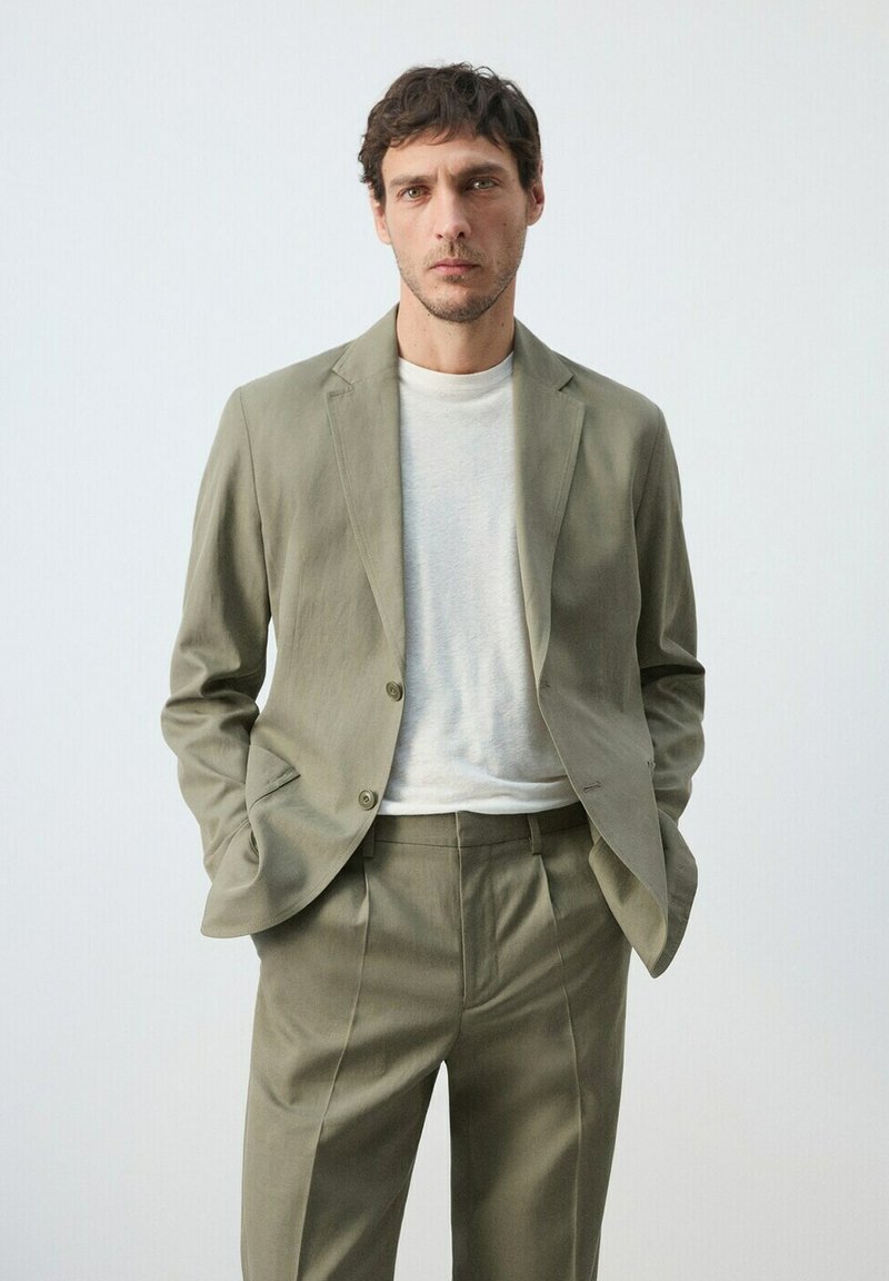 Homme portant un costume vert olive clair et un t-shirt blanc à col rond, debout les mains dans les poches devant un fond clair uni.