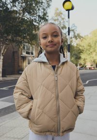 Giacca beige trapuntata con chiusura a zip frontale, cappuccio grigio e polsini a costine. Presenta cuciture a rombi e tasche laterali. Indossata sopra una camicia scura.