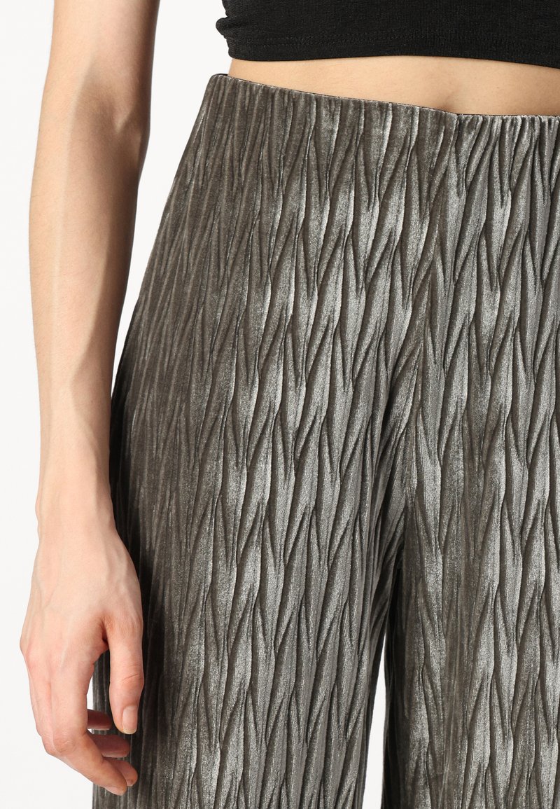 Texturierte graue Hose mit einem vertikalen, wellenförmigen Muster, gefertigt aus einem weichen Material. Hoch tailliertes Design, das Details des Stoffes und der Passform zeigt.