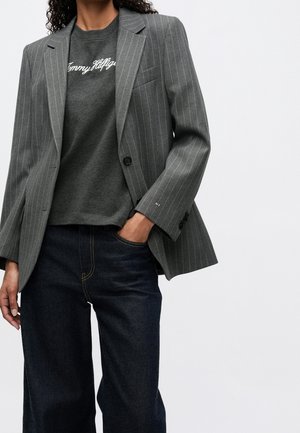 Osoba nosząca szary marynarkę w prążki na ciemnoszarej bluzie "Tommy Hilfiger" i ciemnoniebieskich dżinsach, z jedną ręką w kieszeni.