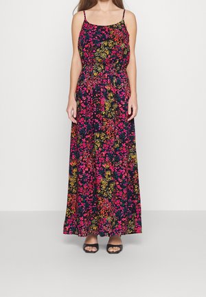 Vrouw draagt een zwarte mouwloze jurk tot op de enkels met roze, gele en rode bloemenpatronen en zwarte sandalen met open tenen.