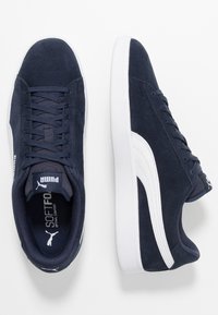 Navy suede sneakers with white accents, featuring a rounded toe, lace-up closure, and padded tongue. Sole is white with textured detailing.