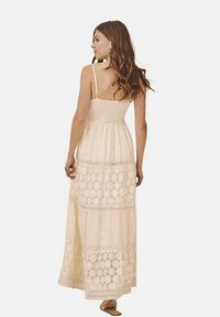 Vestido maxi en beige cremosa, hecho de un tejido ligero. Presenta un corpiño ajustado, tirantes de espagueti ajustables y un detalle de ojales en capas.