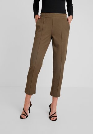 Kobieta ubrana w krótkie oliwkowo-zielone spodnie z przednimi szwami, elastycznym pasem i bocznymi kieszeniami, czarną bluzkę z długim rękawem oraz czarne sandały na wysokim obcasie.