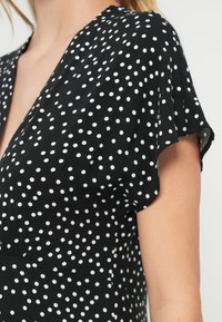 Vestido negro con lunares blancos, con escote en V y mangas cortas. Hecho de material suave con una textura lisa.