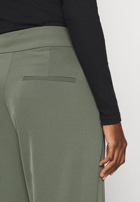 Korkeavyötäröiset, oliivinvihreät leveät housut, joissa on sileä kangas ja takatasku. Musta pitkähiha toppi näkyy osittain.