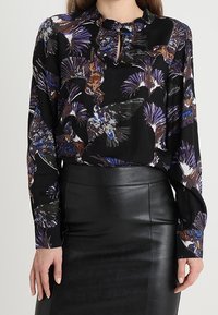 Svart blus med livfullt fågeltryck i blå och bruna nyanser, med långa ärmar, rynkad halsringning och en nyckelhålsutskärning framtill.