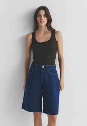 Naine, kes kannab musta lainelise servaga tank-toppi ja lahtisi, põlvepikkusi tumesiniseid teksapükse, taustaks hele ühevärviline pind.