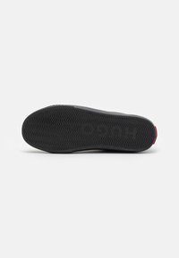 Suela de zapatilla negra con material de goma, patrón en zigzag para tracción y un pequeño acento rojo en el talón.