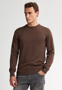 Suéter marrón de manga larga y cuello redondo, hecho de un tejido de punto suave. Los puños y el dobladillo acanalados añaden textura. Combinado con vaqueros grises.
