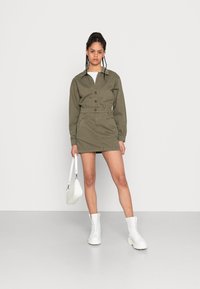 Vestido camisa oversized verde-oliva com botões à frente, cintura elástica e dois bolsos. Combinado com botas de cano curto brancas e uma mala pequena.