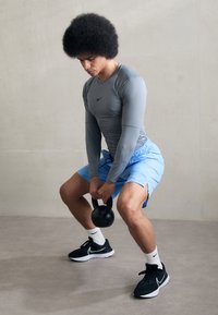 Atletický muž vykonáva zdvih s kettlebellom. Na sebe má šedú dlhú mikinu, modré šortky a čierne športové topánky s bielymi akcentmi.