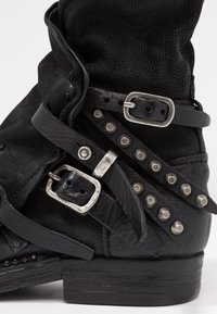 Svart läderstövel med dekorativa spännen, metalldetaljer längs sidan och en strukturerad yta. Staplad klack med robust design.