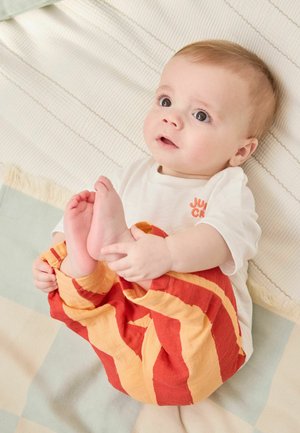 Niemowlę leżące na kocu, ubrane w białą koszulkę i czerwono-pomarańczowe spodnie w paski, trzymające jedną stopę i patrzące w górę szeroko otwartymi oczami.