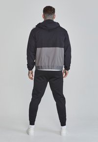 Jachetă color-block în nuanțe de negru și gri, cu glugă, manșete elastice și talie elastică. Asortată cu pantaloni sport negri și adidași albi.