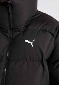 Chaqueta de plumas negra con cuello alto, con cremallera frontal y un logo de Puma en blanco en el pecho. Textura suave, diseño acolchado.