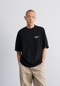 Crna oversized majica s okruglim izrezom, s bijelim izvezenim logom. Model nosi bež hlače, prikazujući opušteni kroj i teksturu tkanine.