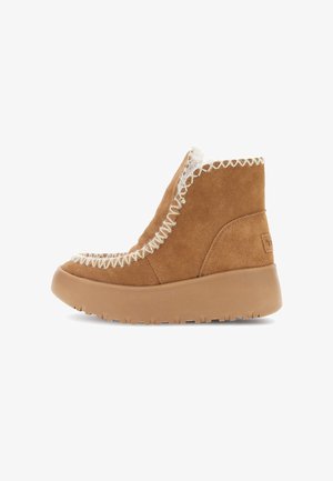 Tan suède enkellaars met witte bontvoering, zigzag witte stiksels en dikke rubberen zool, zijaanzicht op een witte achtergrond.