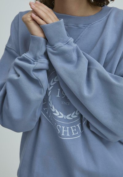 Gaiši zils džemperis ar apaļu kakla izgriezumu, ribu aprocēm un priekšējo grafiku, kurā ir apļveida dizains ar lauru lapām un tekstu.