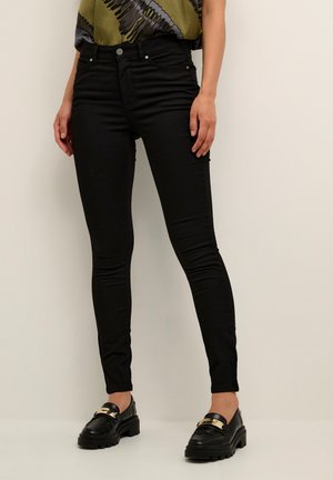 Jean noir slim-fit avec cinq poches, présentant des accessoires en métal argenté et une taille haute. Associé à un haut vert à motifs et des mocassins noirs.