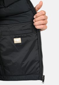 Intérieur d'une veste noire rembourrée montrant une main tenant la fermeture éclair. Présente une étiquette en tissu avec "T-KIT" et une petite poche sur le côté gauche.