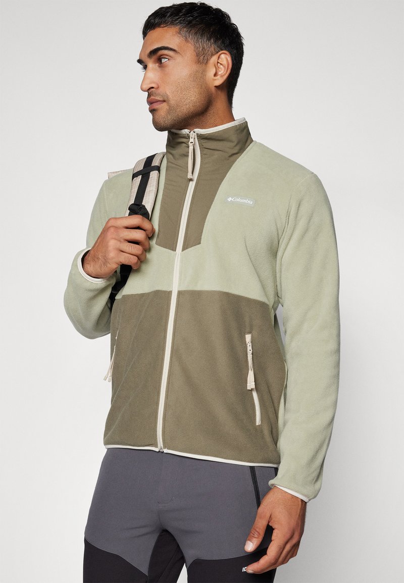 Veste en polaire de couleur vert clair et olive, avec un col haut, des poches zippées et un design de panneau contrasté. Tissu texturé.