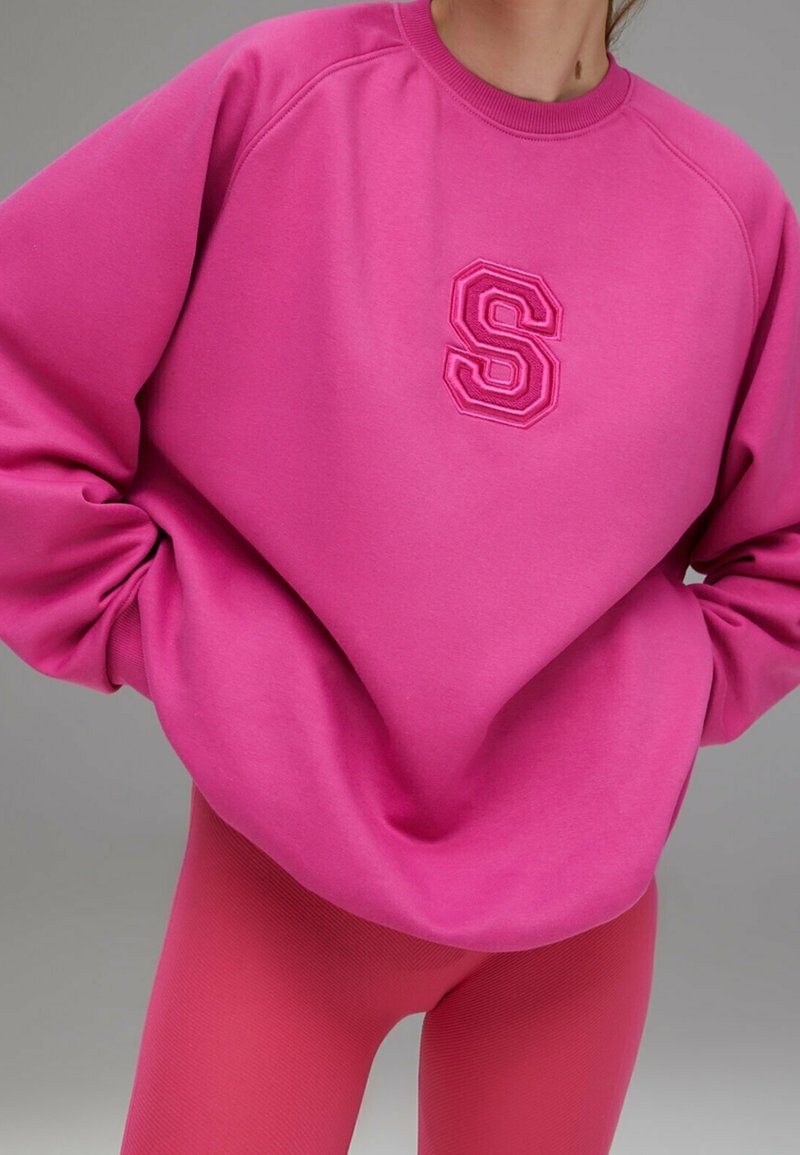 Personne portant un sweat-shirt rose vif avec un emblème "S" en relief et un legging rose assorti, les mains dans les poches du sweat-shirt.